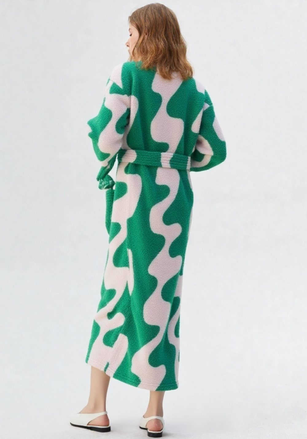 Vue de dos d'une femme en robe de chambre polaire longue verte et rose à motifs abstraits, ceinture, sandales blanches, longueur mollet