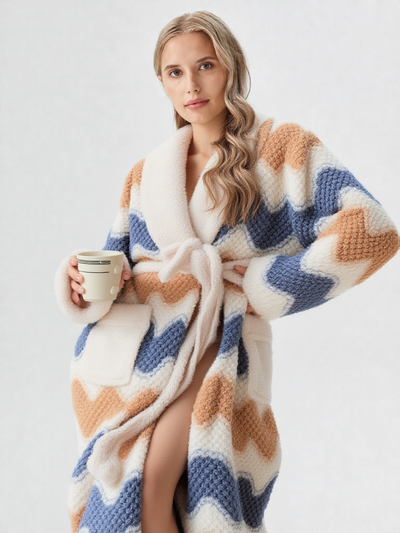 Femme blonde tenant une tasse café, robe de chambre polaire longue à motifs zigzag bleu ardoise et caramel sur fond crème, col châle, ceinture et poches